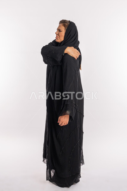 التهاب العضلات والمفاصل، الحاجه للعلاج وطلب المساعده، بورتريه لامرأة خليجيه إماراتية ترتدي العباءة السوداء تشعر بالمرض و التعب، سيدة عربية سعودية تضع يدها على كتفها بإيماءات تدل على الألم الشديد، صورة الجسم بالطول الكامل, خلفية بيضاء
