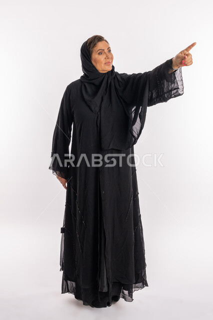 الاشارة إلى مكان ما بحركات الاختيار, امرأة خليجية اماراتية تنظر الى شيء ما بتعابير البشاشة والسرور, بورتريه لسيدة عربية سعودية مبتسمة ترتدي العباءة السوداء ترفع إصبع السبابة إلى الاعلى بتعابير التأكد والتحديد, الجسم بالطول الكامل,  خلفية بيضاء