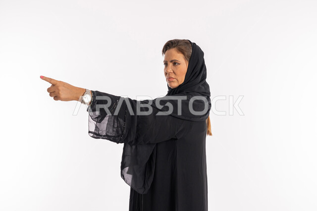 الإتهام و إلقاء اللوم و التوبيخ، طلب التوقف عن فعل الأخطاء، امرأة خليجية اماراتية تشعر بالإنزعاج والرغبة بالإنتقام، بورتريه مقرب من الجانب لسيدة عربية سعودية ترتدي العباءة السوداء ترفع اصبع السبابه لتحديد شخص ما ( أنت )، خلفية بيضاء