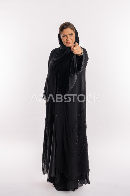 Reprimanding, accusing, and blaming, asking to stop making mistakes, an Emirati Gulf woman feeling annoyed and wanting revenge, a close-up portrait of a Saudi Arab woman wearing a black abaya, raising her index finger forward to identify someone (you), full-length body, white background.