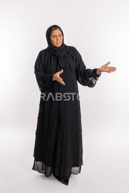 رفع اليدين بحركات تدل على القرف ,بورتريه لسيدة عربيه سعودية محجبة ترتدي العباءة السوداء تنظر الى شيء ما بتعابير الاشمئزاز, امرأة اماراتية خليجية تفتح ذراعيها بايماءات النفور, صورة الجسم بالطول الكامل, خلفية بيضاء