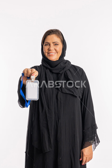 النظر إلى الكاميرا بإيماءات السعادة والسرور، موك اب ابيض، بورتريه لامرأة عربية سعودية مبتسمة ترتدي العباءة السوداء تحمل بيدها بطاقة تعريفية، سيدة خليجية إماراتية تعرض كرت فارغ مستطيل الشكل ,توضيح أهمية الهوية الشخصية, خلفية بيضاء