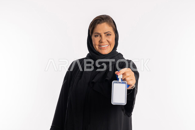 عرض كرت فارغ مستطيل الشكل، موك اب ابيض، بورتريه مقرب لامرأة عربية سعودية مبتسمة ترتدي العباءة السوداء تحمل بيدها  بطاقة تعريفية، سيدة خليجية إماراتية تنظر إلى الكاميرا بإيماءات السعادة والسرور،  توضيح أهمية الهوية الشخصية، خلفية بيضاء