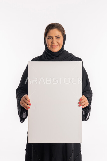 عرض لوحة بيضاء مستطيله فارغة ,وسائل تعليمية و لوحات توضيحية, امرأة اماراتية تنظر الى الكاميرا بتعابير السرور، العروض والإعلانات التسويقية, بورتريه لسيدة عربية خليجية سعودية ترتدي عباءة سوداء تمسك بيديها موك اب مستطيل أبيض, خلفية بيضاء