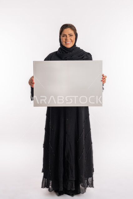 Teaching aids and illustrative panels, an Emirati woman displaying a blank rectangular white board, marketing offers and advertisements, a portrait of a Saudi Arabian Gulf woman wearing a black abaya, holding a white rectangular mock-up in her hands, looking at the camera with an expression of pleasure, full-length body, white background