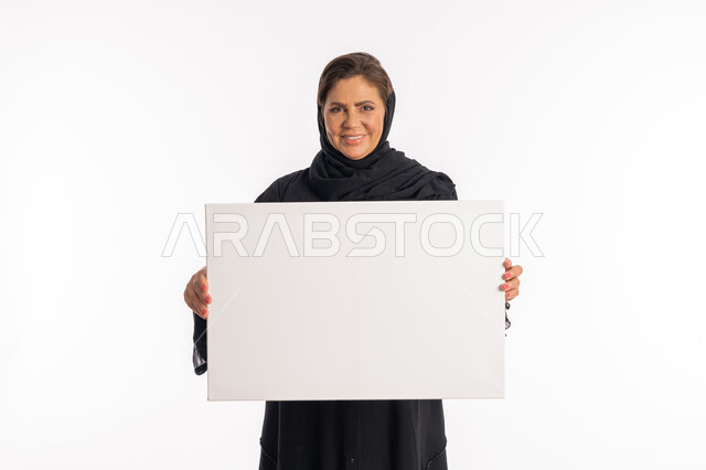 النظر الى الكاميرا بتعابير السرور, لوحات توضيحية ووسائل تعليمية, امرأة اماراتية مبتسمة تعرض لوحة بيضاء مستطيلة فارغة، العروض والإعلانات التسويقية, بورتريه لسيدة عربية خليجية سعودية ترتدي عباءة سوداء تمسك بيديها موك اب مستطيل أبيض, خلفية بيضاء