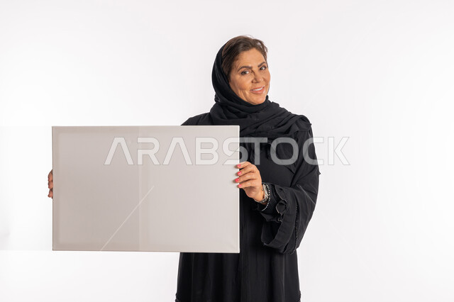 لوحات توضيحية ووسائل تعليمية, امرأة اماراتية مبتسمة تعرض لوحة بيضاء مستطيلة فارغة، العروض والإعلانات التسويقية, بورتريه لسيدة عربية خليجية سعودية ترتدي عباءة سوداء تمسك بيديها موك اب مستطيل أبيض, النظر الى الكاميرا بتعابير السرور, خلفية بيضاء