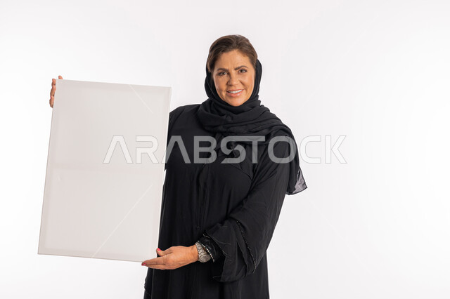 لوحات توضيحية ووسائل تعليمية, امرأة اماراتية مبتسمة تعرض لوحة بيضاء مستطيلة فارغة، العروض والإعلانات التسويقية, بورتريه لسيدة عربية خليجية سعودية ترتدي عباءة سوداء تمسك بيديها موك اب مستطيل أبيض, النظر الى الكاميرا بتعابير السرور, خلفية بيضاء