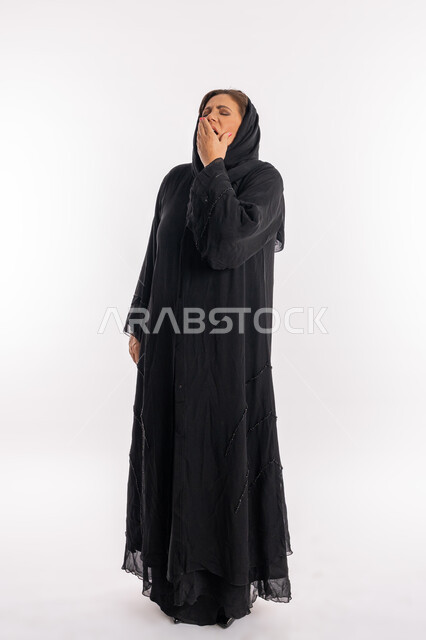 الحاجة إلى النوم, الوقوف بحركات التعب و النعاس, امراه سعودية تشعر بالكسل والخمول وطلب الراحة, بورتريه لسيدة عربيه خليجية اماراتية محجبة ترتدي العباءة السوداء تضع يدها على فمها تقوم بالتثاؤب, السهر لوقت متأخر, صورة الجسم بالطول الكامل, خلفية بيضاء