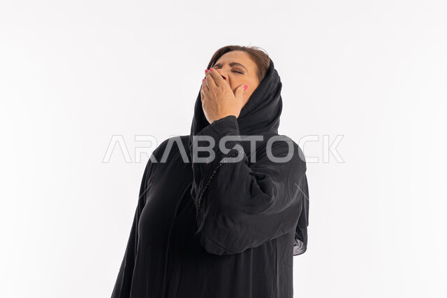 Staying up late, needing to sleep, standing with tired and sleepy movements, a Saudi woman feeling lazy and lethargic and seeking rest, a close-up portrait of a veiled Arab Gulf Emirati woman wearing a black abaya, putting her hand over her mouth and yawning, white background