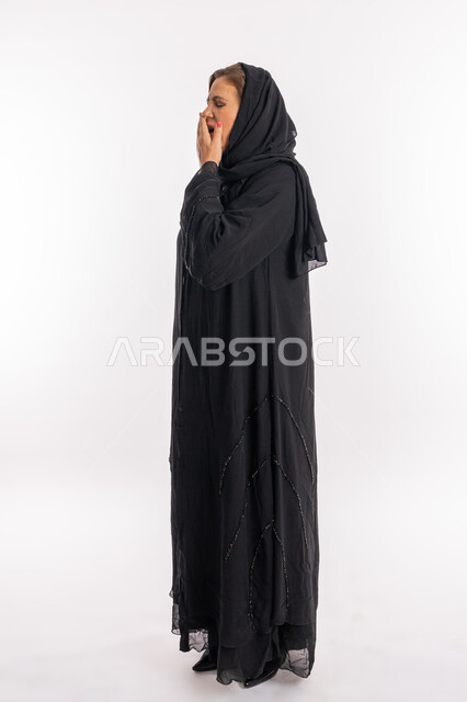الحاجة إلى النوم و النعاس والارهاق, امراه سعودية تشعر بالكسل والخمول وطلب الراحة, بورتريه من الجانب لسيدة عربيه خليجية اماراتية محجبة ترتدي العباءة السوداء تضع يدها على فمها تقوم بالتثاؤب, السهر لوقت متأخر, صورة الجسم بالطول الكامل, خلفية بيضاء