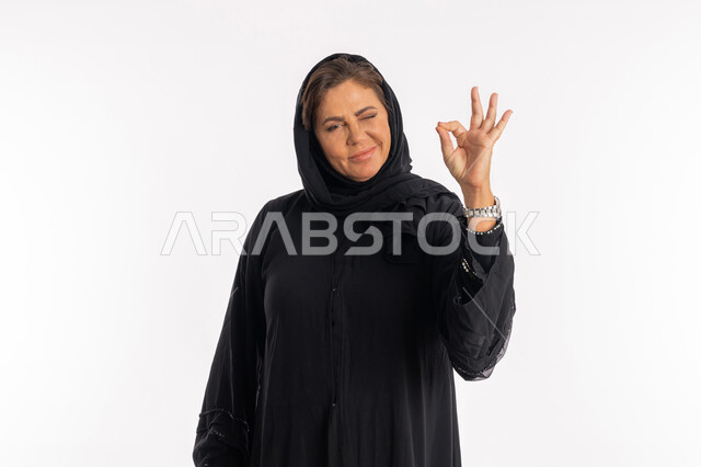 إيماءات القبول و الموافقة, النظر الى الكاميرا بتعابير عين ووجه تدل على الاعجاب, سيدة سعودية تشعر بالرضى، بورتريه مقرب لامراة عربية خليجية اماراتية مبتسمة محجبة ترتدي العباءة السوداء ترفع يدها للاعلى بعلامة الجودة و الامتياز, خلفية بيضاء