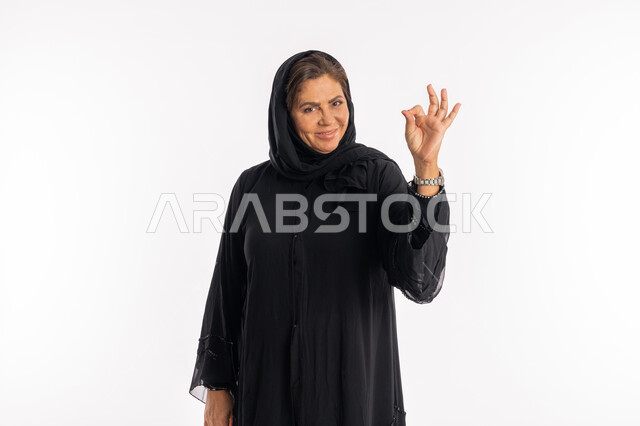 النظر الى الكاميرا بتعابير الاعجاب, سيدة سعودية تشعر بالرضى، بورتريه لامراة عربية خليجية اماراتية مبتسمة محجبة ترتدي العباءة السوداء ترفع يدها للاعلى بإيماءات القبول و الموافقة، علامة الامتياز و الجودة, خلفية بيضاء