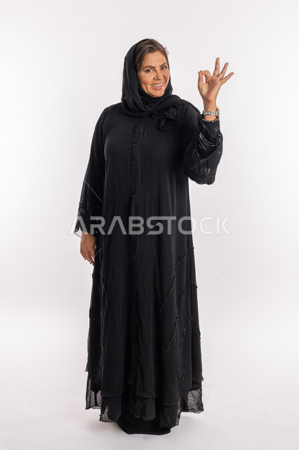علامة الامتياز و الجودة، النظر الى الكاميرا بتعابير السرور, سيدة سعودية تشعر بالإعجاب والرضى، بورتريه لامراة عربية خليجية اماراتية مبتسمة محجبة ترتدي العباءة السوداء ترفع يدها  للاعلى بإيماءات القبول و الموافقة، صورة الجسم بالطول الكامل, خلفية بيضاء