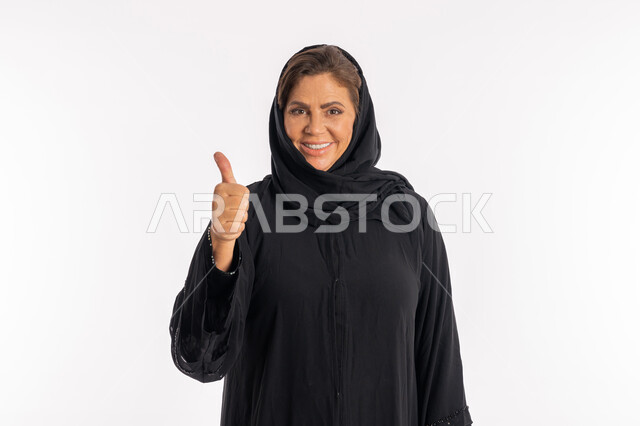 تعابير الموافقة و القبول ,النظر الى الكاميرا بإيماءات السرور، سيدة سعودية تشعر بالإعجاب والرضى، بورتريه مقرب لامراة عربية خليجية اماراتية مبتسمة محجبة ترتدي العباءة السوداء ترفع اصبع الابهام للاعلى بعلامة الامتياز و الجودة, خلفية بيضاء