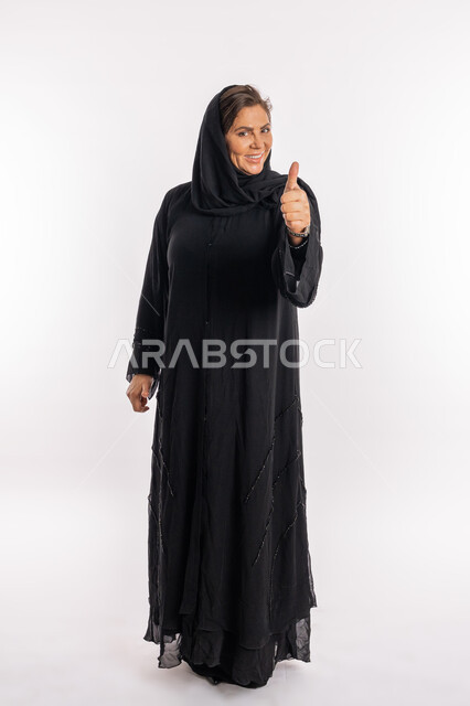 إيماءات الموافقة و القبول ,النظر الى الكاميرا بتعابير السرور، سيدة سعودية تشعر بالإعجاب والرضى، بورتريه لامراة عربية خليجية اماراتية مبتسمة محجبة ترتدي العباءة السوداء ترفع اصبع الابهام للاعلى بعلامة الامتياز و الجودة، صورة الجسم بالطول الكامل, خلفية بيضاء