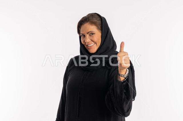 إيماءات القبول و الموافقة ,النظر الى الكاميرا بتعابير السرور، سيدة سعودية تشعر بالإعجاب والرضى، بورتريه مقرب لامراة عربية خليجية اماراتية مبتسمة محجبة ترتدي العباءة السوداء ترفع اصبع الابهام للاعلى بعلامة الامتياز و الجودة، خلفية بيضاء