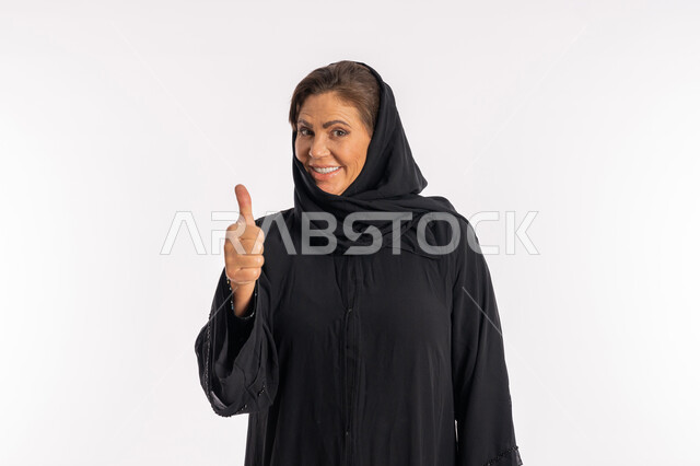 النظر الى الكاميرا بتعابير السرور, علامة الامتياز و الجودة، سيدة سعودية تشعر بالإعجاب والرضى، بورتريه مقرب لامراة عربية خليجية اماراتية مبتسمة محجبة ترتدي العباءة السوداء ترفع اصبع الابهام للاعلى بإيماءات القبول و الموافقة، خلفية بيضاء