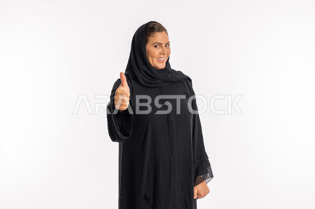 علامة الامتياز و الجودة، النظر الى الكاميرا بتعابير السرور, سيدة سعودية تشعر بالإعجاب والرضى، بورتريه لامراة عربية خليجية اماراتية مبتسمة محجبة ترتدي العباءة السوداء ترفع اصبع الابهام للاعلى بإيماءات القبول و الموافقة، خلفية بيضاء