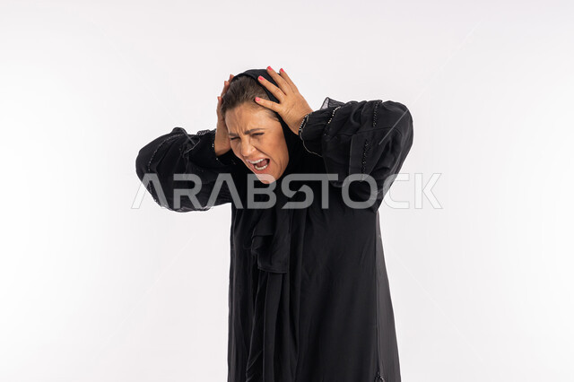 الحياه الفاشله و الامراض النفسيه ،بورتريه مقرب لامرأة عربيه خليجية اماراتية محجبة ترتدي العباءة السوداء تضع يديها على رأسها بايماءات الغضب و العصبية و الاثارة, تعابير الصراخ الشديد, سيدة سعودية تشعر بالانزعاج و الرغبة بالانتقام, خلفية بيضاء