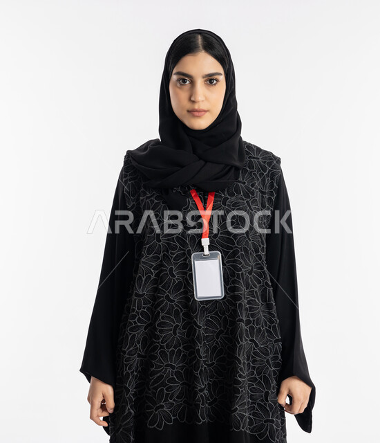 النظر إلى الكاميرا بثقة وجدية, الملامح العربية القوية, ارتداء بادج شخصي, التعبير عن الذات, بورتريه لشابة عربية خليجية إماراتية ترتدي العباءة تقف بتعابير القوة, سيدة سعودية تضع هوية شخصية, خلفية بيضاء
