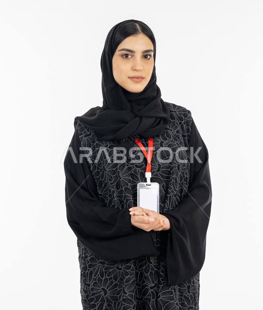 ارتداء هوية شخصية, الوقوف باستقامة بإيماءات الجدية والثقة بالنفس, بورتريه لإمرأة عربية خليجية إماراتية ترتدي العباءة تضع بادج شخصي بشريط أحمر, سيدة سعودية تنظر إلى الكاميرا, أناقة النساء الخليجيات, الاهتمام بالمظهر الخارجي, خلفية بيضاء