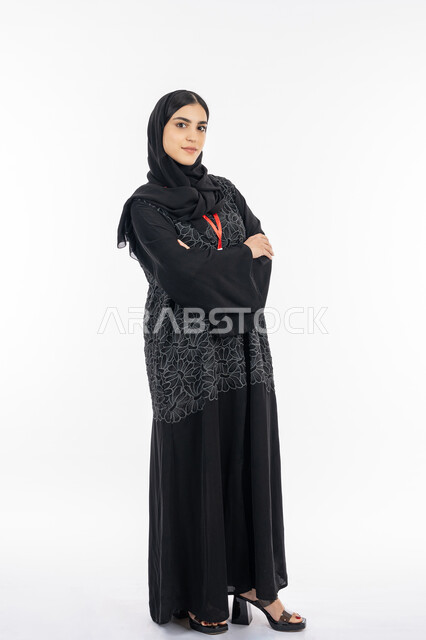 النعومة والأنوثة والاهتمام بالمظهر الخارجي, بورتريه لشابة عربية خليجية إماراتية ترتدي العباءة تقف بإيماءات ايدي متقاطعة, سيدة سعودية مبتسمة تنظر إلى الكاميرا بثقة, أناقة النساء الخليجيات, صورة الجسم بالطول الكامل, خلفية بيضاء