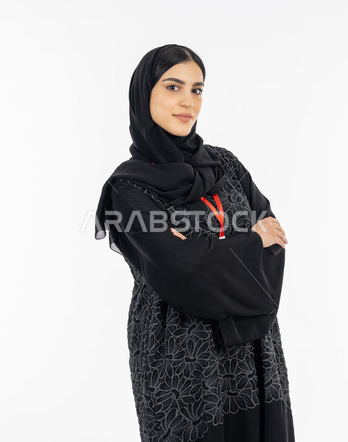 Femininity, softness, and attention to external appearance, elegance of Gulf women, portrait of an Arab Gulf Emirati young woman wearing an abaya, standing with crossed hand gestures, a smiling Saudi woman looking at the camera confidently, white background