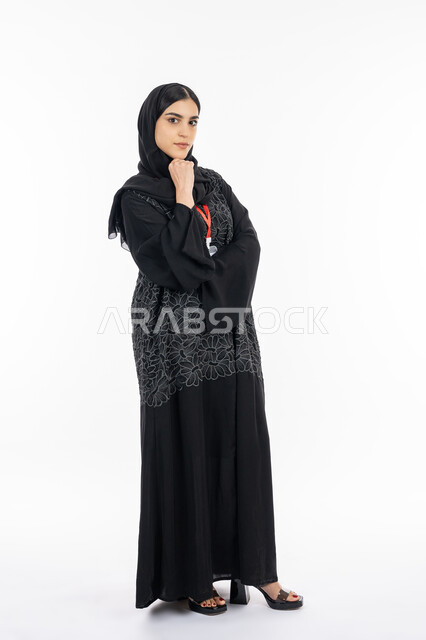 مفهوم الأنوثة والنعومة واللطافة, الملامح العربية القوية, بورتريه لشابة عربية خليجية إماراتية ترتدي العباءة تنظر إلى الكاميرا, سيدة سعودية تضع يدها على ذقنها بإيماءات التفكير, صورة الجسم بالطول الكامل, خلفية بيضاء