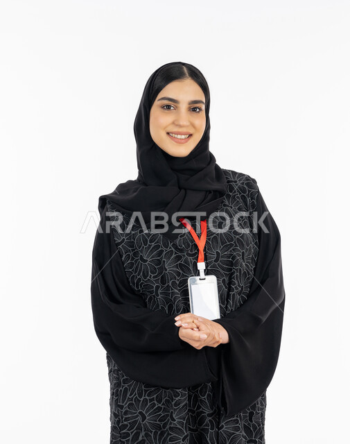 النظر إلى الكاميرا بتعابير السرور, بورتريه لشابة عربية خليجية إماراتية ترتدي العباءة تضع بادج شخصي بشريط أحمر, سيدة سعودية تقف بابتسامة, الهوية الشخصية والتعبير عن الذات، بطاقة تعريفية للطلاب والموظفين, خلفية بيضاء