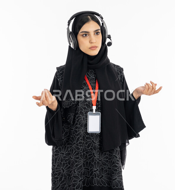 Using headphone technology in remote speaking programs, answering customers’ questions, standing in confusion, astonishment and questioning, a portrait of a Saudi Gulf Arab woman wearing an abaya who works in the field of customer service, an Emirati woman wearing a personal badge, a white background.