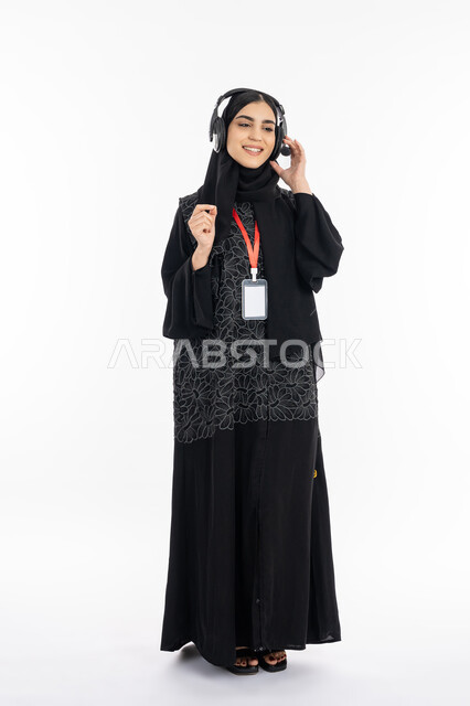 Wearing an identification card, working in the technical support department, using headphone technology, a portrait of an Arab Gulf Emirati young woman wearing an abaya who works in the field of customer service, a Saudi woman putting her hands on the headset to listen and focus on the customer’s questions, full body image, white background
