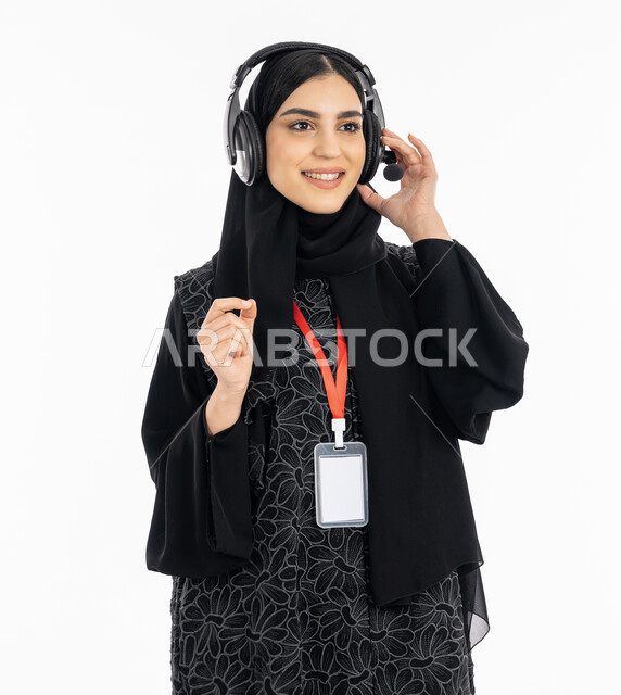 النظر إلى شيء ما بسرور, العمل في قسم الدعم الفني, استخدام تقنية الهيدفون, ارتداء بادج شخصي بورتريه لشابة عربية خليجية إماراتية ترتدي العباءة تعمل في مجال خدمة العملاء, سيدة سعودية تضع يديها على سماعة الرأس للإصغاء والتركيز على اسئلة العميل, خلفية بيضاء