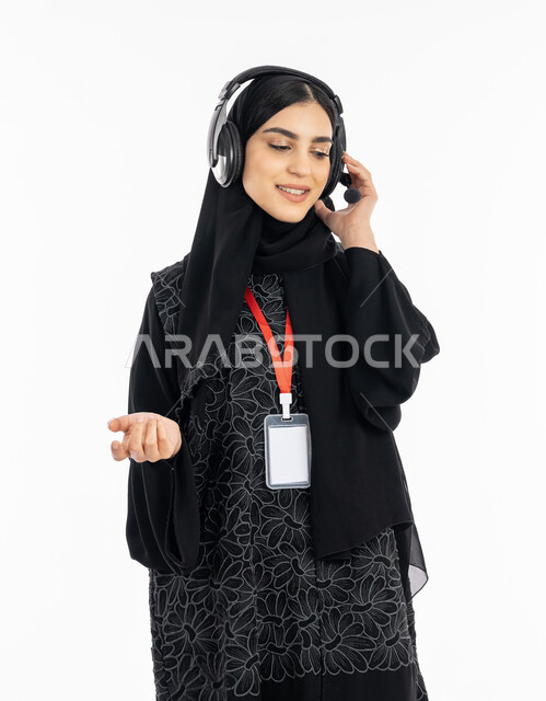 استخدام تقنية الهيدفون, الرد على اسئلة الزبائن ببشاشة, ارتداء بادج شخصي, بورتريه لشابة عربية خليجية إماراتية ترتدي العباءة تعمل في مجال خدمة العملاء, سيدة سعودية تضع يديها على سماعة الرأس للإصغاء للعميل, العمل في قسم الدعم الفني, خلفية بيضاء