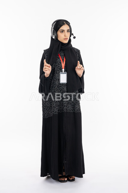 الرد على اسئلة الزبائن, الوقوف بدهشة واستغراب وصدمة, بورتريه لإمرأة عربية خليجية سعودية ترتدي العباءة تعمل في مجال خدمة العملاء, سيدة اماراتية تضع بادج شخصي, استخدام تقنية الهيدفون في برامج التحدث عن بعد, صورة الجسم بالطول الكامل, خلفية بيضاء