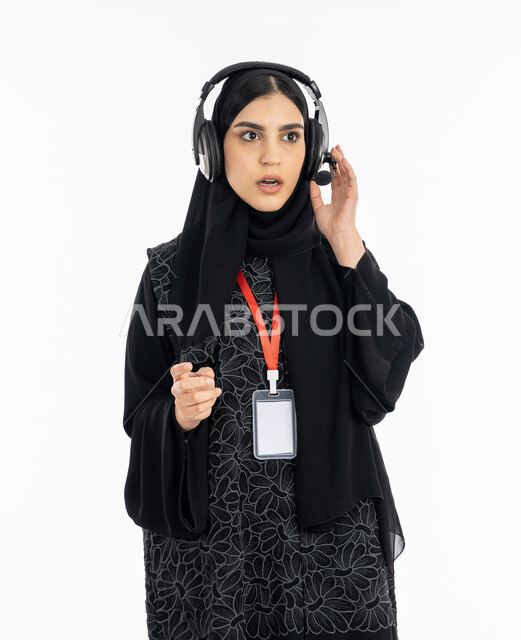 Standing in astonishment, astonishment and shock, a portrait of a Saudi Gulf Arab woman wearing an abaya working in the field of customer service, an Emirati woman wearing a personal badge, using headphone technology in long-distance programs, answering customers’ questions, white background