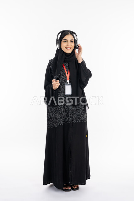 الرد على اسئلة الزبائن ببشاشة, استخدام تقنية الهيدفون, ارتداء بادج شخصي, بورتريه لشابة عربية خليجية إماراتية ترتدي العباءة تعمل في مجال خدمة العملاء, سيدة سعودية تقف باستقامة بتعابير السعادة, صورة الجسم بالطول الكامل, خلفية بيضاء