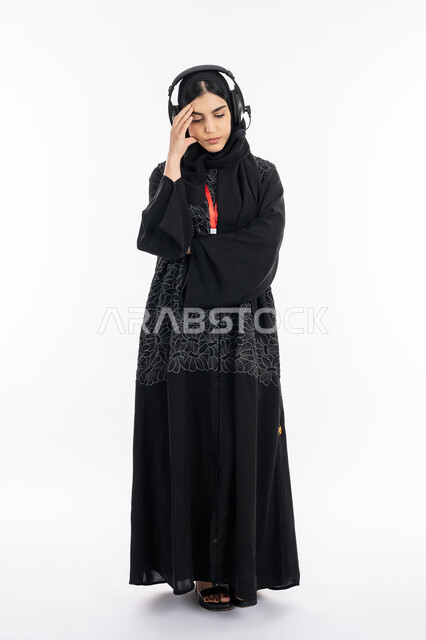 التعب والملل والحزن, الشعور بألم الرأس, بورتريه لشابة عربية خليجية إماراتية ترتدي العباءة تضع يدها على رأسها, سيدة سعودية تقف بإيماءات عدم الراحة, العمل في مجال خدمة العملاء, استخدام تقنية الهيدفون، برامج التحدث عن بعد, صورة الجسم بالكامل، خلفية بيضاء