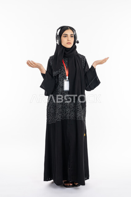 الوقوف ورفع اليدين بإيماءات الحيرة وعدم الرضا والقبول, بورتريه لشابة عربية خليجية إماراتية ترتدي العباءة تستخدم تقنية الهيدفون, سيسدة سعودية تضع بادج شخصي, العمل في مجال خدمة العملاء، برامج التحدث عن بعد، صورة الجسم بالكامل, خلفية بيضاء