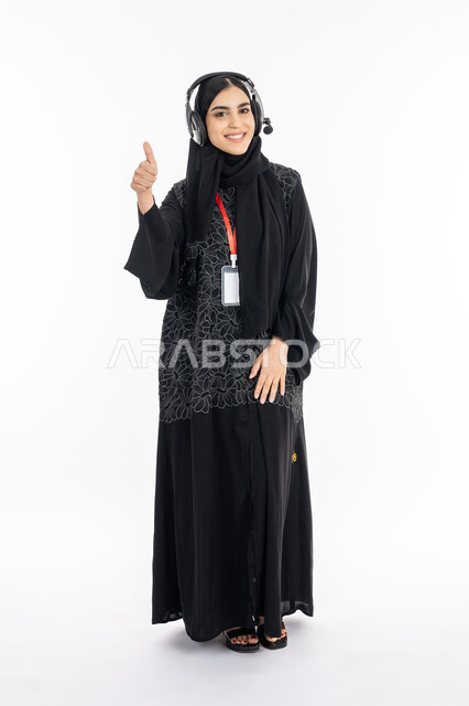 Answering inquiries in the technical support section, integrating work with technology and technology in practical life, a Saudi woman wearing a headphone, a portrait of a smiling Arab Gulf Emirati woman wearing an abaya, raising her finger with the sign of quality and excellence, full body photo, white background