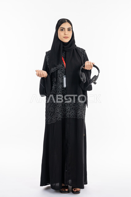 النظر إلى الكاميرا والوقوف باستقامة, بورتريه لشابة عربية خليجية إماراتية ترتدي العباءة تسخدم تقنية الهيدفون, سيدة سعودية مبتسمة تقضي وقت ممتع بسماع الموسيقى, استخدام التقنيات الحديثة والمتطورة, صورة الجسم بالطول الكامل, خلفية بيضاء