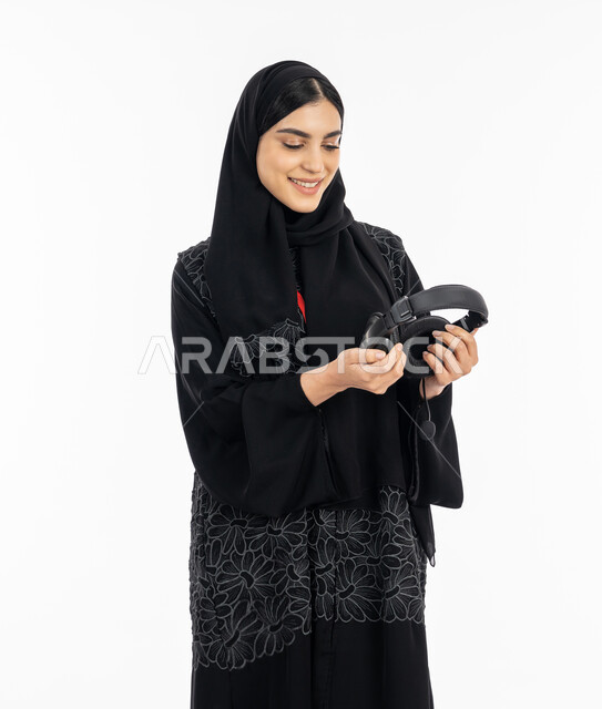 التسيلة والترفيه وقضاء وقت الفراغ بالأنشطة الممتعه, الاستمتاع بسماع الموسيقى,  بورتريه لإمرأة عربية خليجية اماراتية ترتدي العباءة تمسك بيدها سماعة الرأس الصوتية, سيدة سعودية مبتسمة تستخدم تقنية الهيدفون, خلفية بيضاء