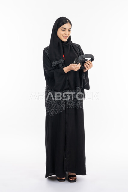 قضاء وقت الفراغ بالأنشطة الممتعه, دمج التقنية الحديثة بالتسلية والترفيه, الاستمتاع بسماع الموسيقى, بورتريه لإمرأة عربية خليجية اماراتية ترتدي العباءة تمسك بيدها سماعة الرأس الصوتية, سيدة سعودية مبتسمة تستخدم تقنية الهيدفون, صورة الجسم بالكامل, خلفية بيضاء