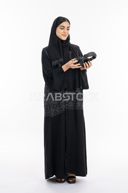 الوقوف باستقامة بإيماءات السعادة, الاستمتاع بسماع الموسيقى, قضاء وقت الفراغ بالأنشطة الترفيهية الممتعه, بورتريه لإمرأة عربية خليجية اماراتية ترتدي العباءة تمسك بيدها سماعة الرأس الصوتية, سيدة سعودية مبتسمة تستخدم تقنية الهيدفون, صورة الجسم بالكامل, خلفية بيضاء