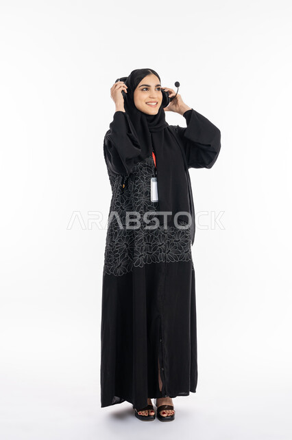 الاندماج والنظر إلى شيء ما بسرور, سيدة سعودية تقوم بالرد على استفسارات الزبائن، ارتداء بادج شخصي بشريط أحمر, تقديم البرامج الاذاعية، بورتريه لإمرأة عربية خليجية اماراتية ترتدي العباءة تضع الهيدفون، العمل في مجال خدمة العملاء، صورة الجسم بالكامل, خلفية بيضاء