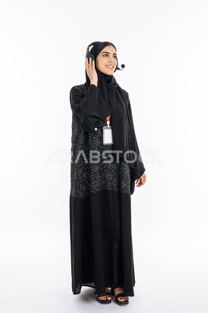 Using modern and advanced technologies, looking at something with gestures of joy and joy, a portrait of an Arab Gulf Emirati young woman wearing an abaya wearing a personal badge with a red ribbon, a smiling Saudi woman placing her headphone in interaction and immersion in listening to music, full-length body portrait, white background