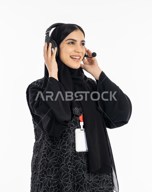 النظر إلى شيء ما بإيماءات البشاشة والبهجة, بورتريه لشابة عربية خليجية إماراتية ترتدي العباءة تعمل في مجال خدمة العملاء, سيدة سعودية مبتسمة تضع الهيدفون بتفاعل واندماج بسماع الموسيقى, استخدام التقنيات الحديثة والمتطورة,  خلفية بيضاء