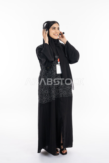 الوقوف والنظر إلى شيء ما بإيماءات البشاشة والبهجة, بورتريه لشابة عربية خليجية إماراتية ترتدي العباءة تعمل في مجال خدمة العملاء, سيدة سعودية تضع الهيدفون بتفاعل واندماج بسماع الموسيقى, استخدام التقنيات الحديثة والمتطورة, صورة الجسم بالطول الكامل, خلفية بيضاء