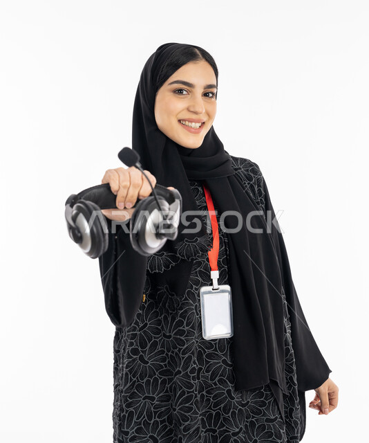 الرد والاجابة على اسئلة الزبائن بإيماءات البشاشة والبهجة, بورتريه لموظفة عربية خليجية إماراتية ترتدي العباءة تعمل في مجال خدمة العملاء, سيدة سعودية تمسك سماعة الرأس, استخدام التقنيات الحديثة في برامج التحدث عن بعد, خلفية بيضاء