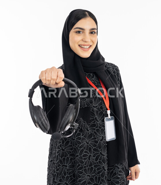 مفهوم خدمة العملاء, استخدام تقنية الهيدفون, العمل في مجال التوزيع الموسيقي, سيدة سعودية تعمل في تقديم البرامج الاذاعية, بورتريه لإمرأة عربية خليجية إماراتية مبتسمة ترتدي العباءة السوداء تضع بطاقة تعريفية شخصية للموظفين وتعرض سماعة الرأس, خلفية بيضاء